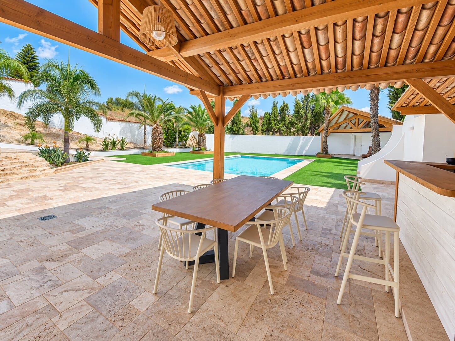 Mesa larga bajo la pérgola de madera, junto a la piscina