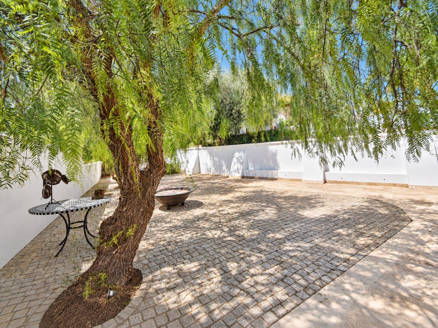 Patio empedrado a la sombra de un gran árbol, junto a los muros encalados